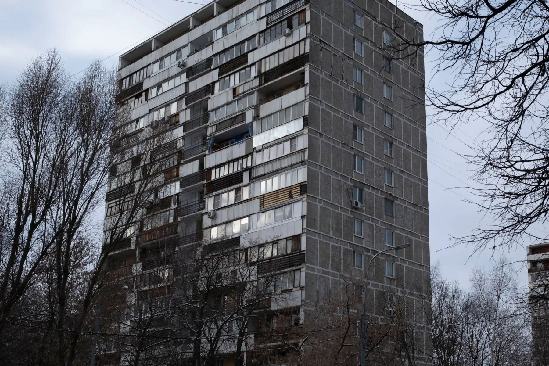 Pilkas daugiaaukštis gyvenamasis namas su balkonais ir medžiais priekyje