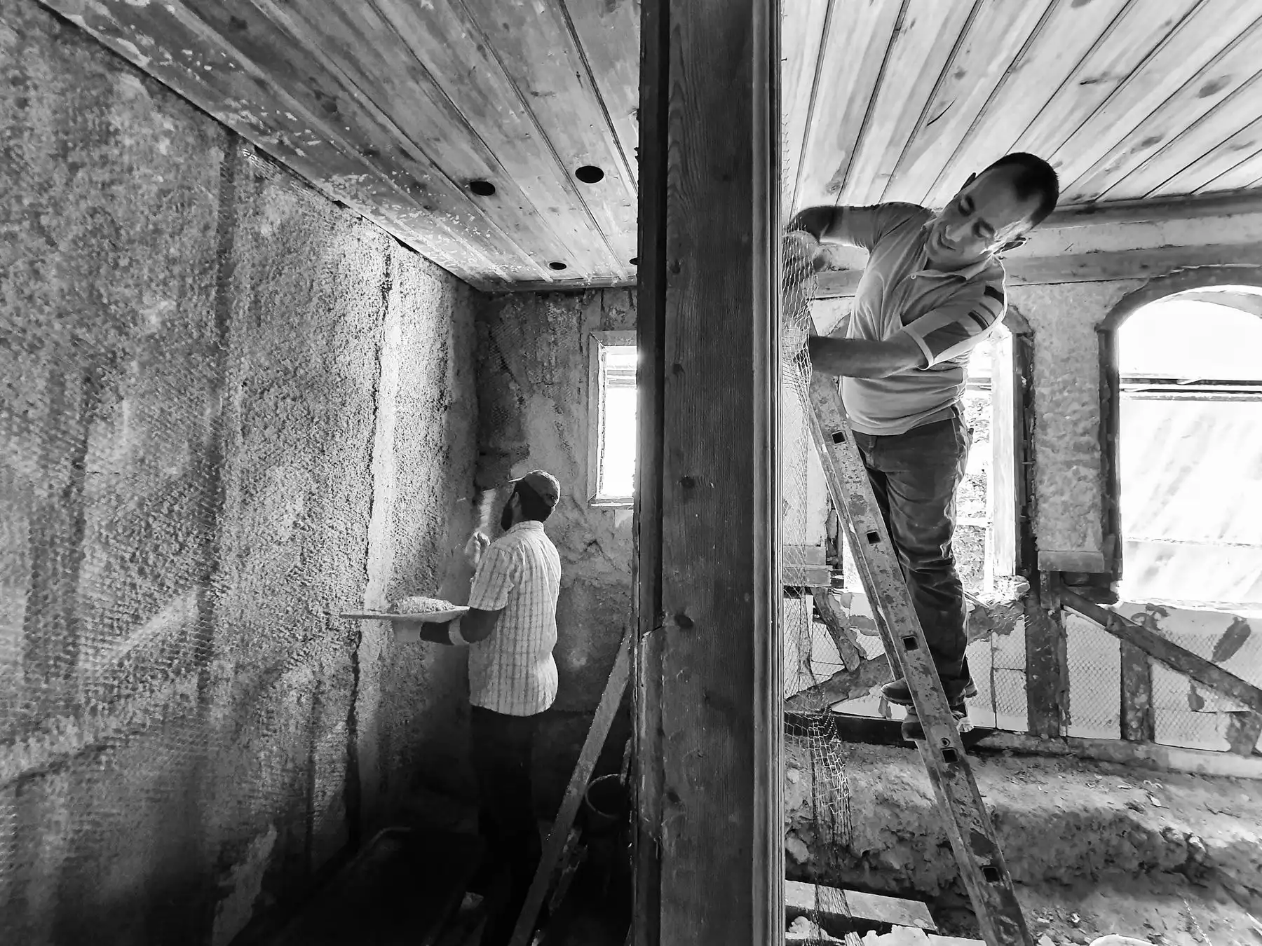 Du meistrai remontuoja seną butą, vienas ant kopėčių tinkuoja sieną